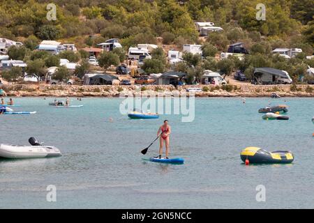 Kosirina, Murter, Croatie - 24 août 2021 : bateaux gonflables amarrés dans une baie calme et des personnes pagayant sur des planches debout, sur une plage de camping Banque D'Images