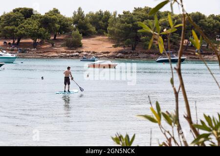 Kosirina, Murter, Croatie - 24 août 2021 : les gens pagayent sur des planches debout et nagent et des bateaux amarrés dans une baie calme et une plage de pins Banque D'Images