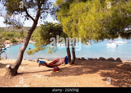 Kosirina, Murter, Croatie - 24 août 2021 : personne se trouvant dans un hamac coloré à l'ombre des pins sur la plage et des bateaux amarrés dans une baie en per Banque D'Images