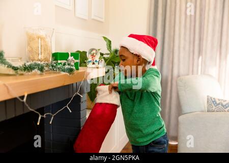 Un garçon afro-américain portant un chapeau de père noël et recherchant un cadeau dans le bas de noël Banque D'Images