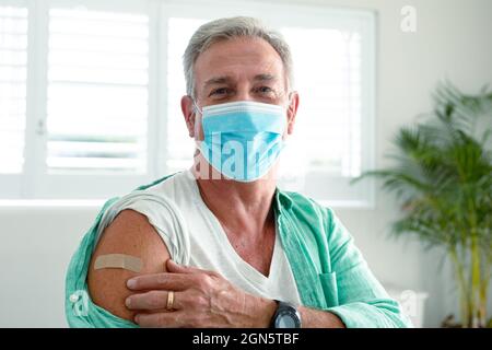 Homme âgé caucasien souriant avec du plâtre sur le bras après la vaccination, portant un masque facial Banque D'Images