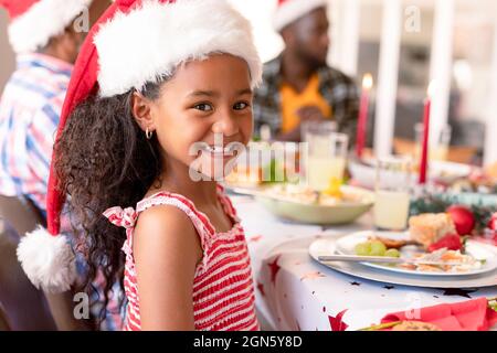 Bonne fille afro-américaine portant un chapeau de père noël assis à la table de noël Banque D'Images