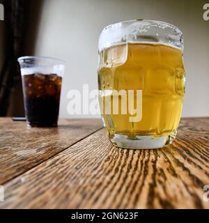 Tasse à bière avec bière pression sur une table rustique avec un verre de boisson au cola en arrière-plan ; désaltérant par temps chaud. Banque D'Images