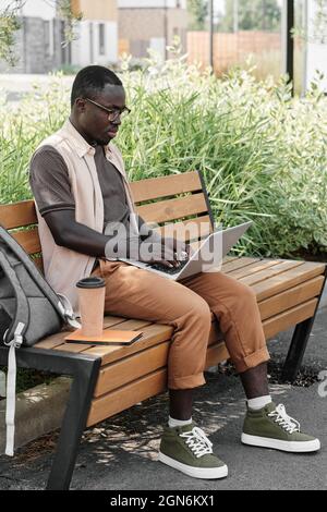 Prise de vue verticale du corps d'un jeune Afro-américain portant des vêtements décontractés assis sur un banc à l'extérieur, travaillant sur un ordinateur portable lors d'une chaude journée d'été Banque D'Images