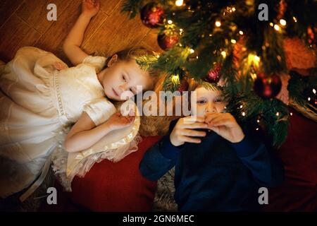 Frère et sœur se trouve sur la moquette près de l'arbre du nouvel an dans un intérieur de noël confortable avec guirlande festive. Mise au point douce sélective, effet de grain de film Banque D'Images