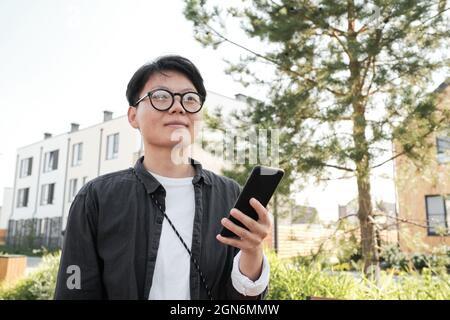 Portrait horizontal bas angle moyen de la jeune femme asiatique avec des cheveux courts portant des lunettes de vue passant du temps à l'extérieur à marcher et à écouter des messages vocaux sur un smartphone Banque D'Images