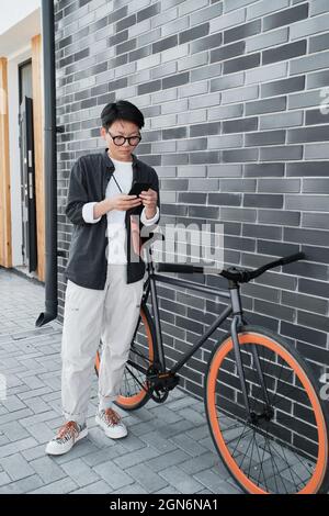 Coupe verticale longue de la jeune femme asiatique aux cheveux courts portant un ensemble élégant et décontracté debout avec un vélo Banque D'Images