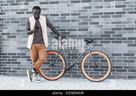 Plan horizontal long du jeune Noir homme portant des vêtements décontractés debout avec un vélo contre un bâtiment en brique grise regardant quelque chose sur un smartphone, espace de copie Banque D'Images