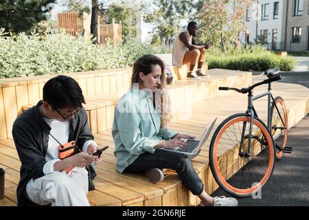 Groupe de trois jeunes gens multiethniques passent la journée d'été dehors assis dans un parc urbain moderne travaillant à distance ou surfant sur Internet sur leurs gadgets Banque D'Images