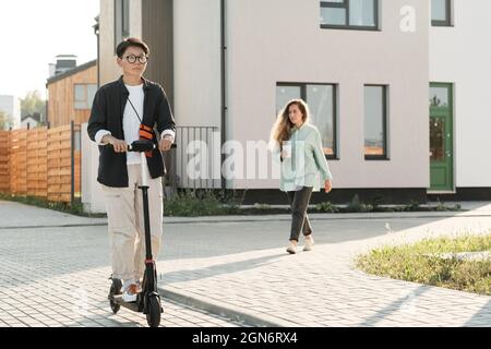 Plan horizontal long de jeune femme asiatique élégante portant des lunettes de vue à cheval e-scooter en plein air chaud et ensoleillé jour d'été Banque D'Images