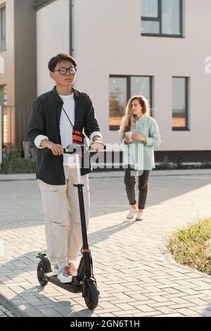 Vue verticale longue d'une jeune femme asiatique sérieuse portant une tenue élégante et des lunettes de vue à cheval sur e-scooter le long de la rue par une chaude journée ensoleillée d'été Banque D'Images