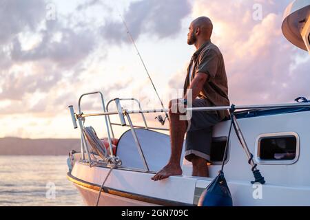 Jeune afro-américain debout avec une canne à pêche sur un voilier pêchant en pleine mer au coucher du soleil Banque D'Images