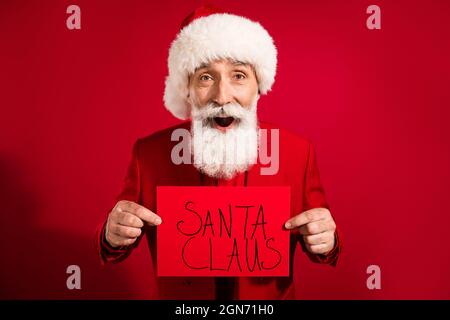 Photo d'un charmant homme mature impressionné habillé costume de noël chapeau tenant l'écriteau du père noël isolé rouge arrière-plan Banque D'Images