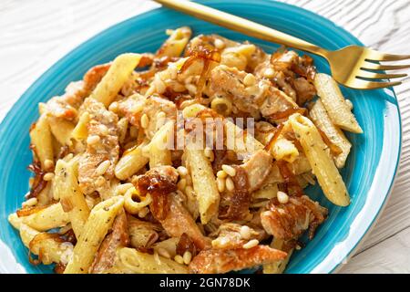 gros plan du poulet alfredo pasta penne avec oignon caramélisé arrosé de pignons de pin et parmesan râpé sur une assiette bleue sur une patte en bois blanc Banque D'Images