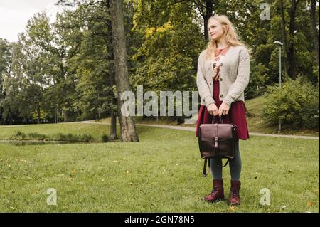 Jeune femme en veste tricotée, collants gris et jupe. Chaussures bordeaux élégantes. Sac en cuir dans les bras. Herbe verte. Mode d'automne. Banque D'Images