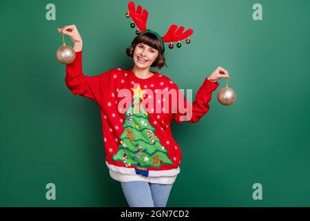 Photo d'une jeune femme adorable vêtue rouge pull-over bois souriant tenant des boules nouvel an isolé couleur vert fond Banque D'Images