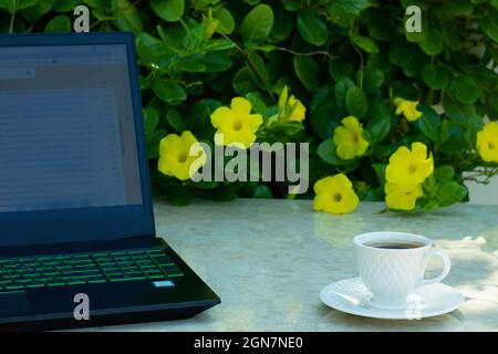 Magnifique tasse à café sur la table en marbre et ordinateur portable sur la gauche de la table. Le fond est un magnifique Bush de fleurs jaunes et de feuilles vertes fraîches. Banque D'Images