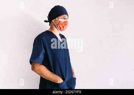 Infirmière, médecin ou vétérinaire de sexe masculin portant un masque rouge et un uniforme bleu foncé à l'hôpital, mains dans les poches, souriant. Médecine, hôpital et soins de santé Banque D'Images