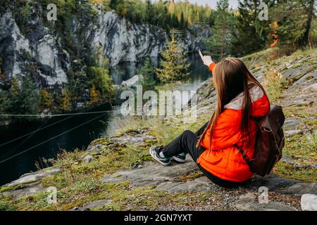 Une jeune fille touriste dans une veste rouge regarde le lac de montagne et pointe sa main sur le côté dans Marble Canyon. Automne à Carélie, Russie. Banque D'Images