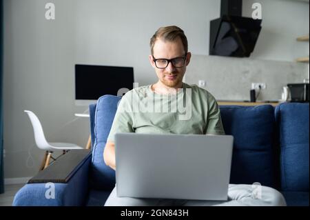 Travailleur indépendant intelligent utilisant un ordinateur portable pour travailler à distance depuis la maison. Un beau gars barbu surfer sur le Web, envoie et répond aux e-mails, développe le logiciel sittin sur le canapé à la maison. L'homme intelligent travaille à distance Banque D'Images