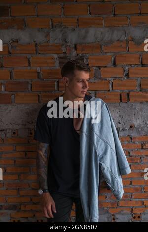 Portrait d'un jeune homme caucasien beau avec un aspect pensif. Portant des vêtements décontractés, regardant de côté, tenant la veste en denim mur de brique rouge sur le fond Banque D'Images
