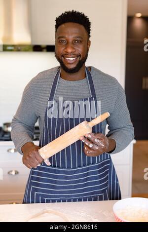 Portrait d'un homme afro-américain heureux portant un tablier, tenant une goupille dans la cuisine Banque D'Images