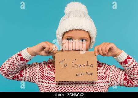 enfant positif en bonnet tricoté tenant la lettre au père noël près du visage isolé sur bleu Banque D'Images