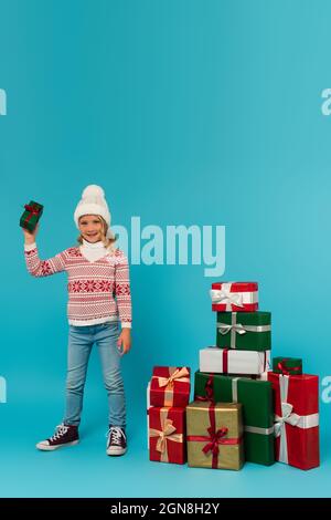 enfant dans un chapeau chaud, un chandail et un jean tenant le présent près de la pile de boîtes-cadeaux sur le bleu Banque D'Images