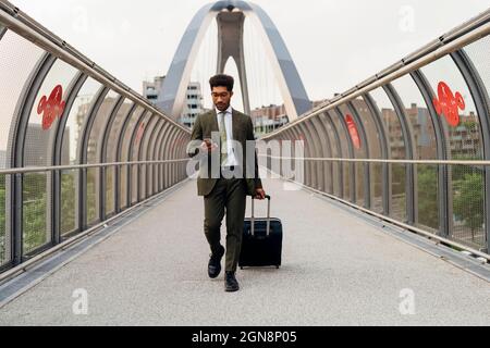 Jeune homme d'affaires utilisant un téléphone portable tout en marchant avec des bagages à roulettes sur le pont Banque D'Images