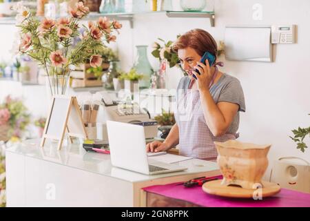 Femme d'entrepreneur mature qui parle sur un téléphone intelligent lorsqu'elle se tient au check-out en magasin Banque D'Images