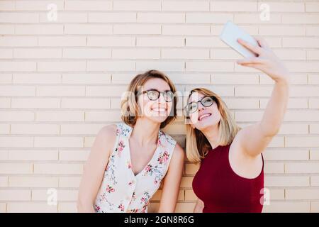 Des femmes souriantes emportant le selfie à travers un smartphone devant un mur de briques Banque D'Images