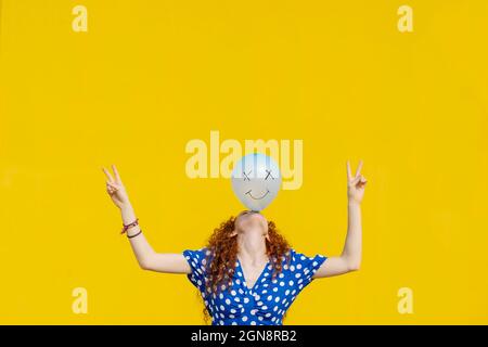 Femme insouciante ballon d'équilibrage avec visage de smiley anthropomorphique devant le mur jaune Banque D'Images