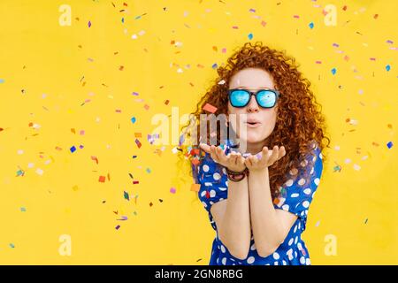 Belle femme soufflant confetti devant le mur jaune Banque D'Images