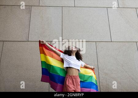 Femme tenant le drapeau arc-en-ciel en se tenant devant le mur Banque D'Images