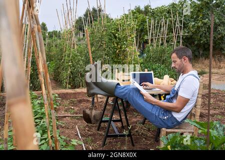 Un agriculteur de sexe masculin lit le presse-papiers tout en étant assis avec les pieds à la table dans le champ agricole Banque D'Images