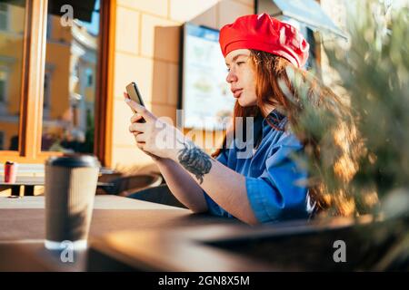 Jeune femme en chapeau de béret utilisant un smartphone au café-terrasse Banque D'Images