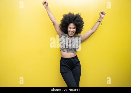 Jeune femme afro excitée avec les bras levés devant le mur jaune Banque D'Images