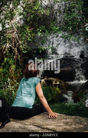 Femme adulte de taille moyenne regardant la chute d'eau tout en étant assise avec un chien en forêt Banque D'Images