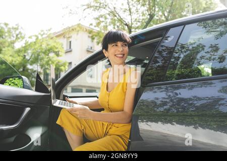 Femme d'affaires souriante assise avec un ordinateur portable en voiture Banque D'Images