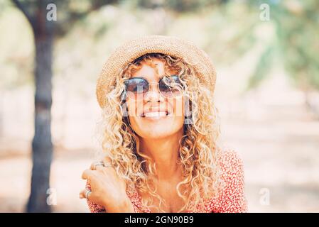 Femme mûre souriante aux cheveux blonds bouclés portant des lunettes de soleil et un chapeau Banque D'Images