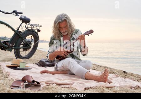 Homme mature avec cheveux gris jouant ukulele à la plage Banque D'Images