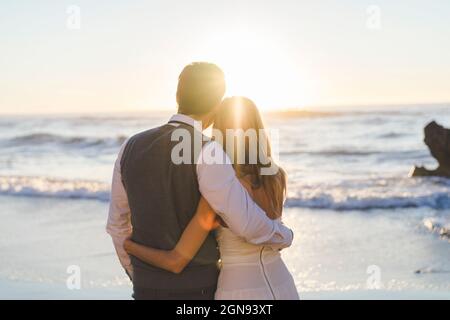 Jeune couple nouvellement mariés avec bras autour de regarder le lever du soleil vue sur la plage Banque D'Images