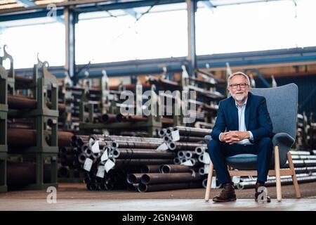 Homme d'affaires souriant assis avec les mains clastées sur un fauteuil en usine Banque D'Images