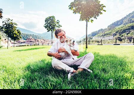 Homme adulte de taille moyenne avec chien d'animal de compagnie assis à pieds croisés sur l'herbe Banque D'Images