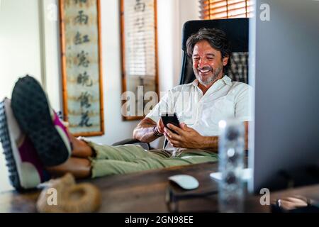 Souriant homme professionnel utilisant le téléphone mobile tout en se relaxant sur une chaise au bureau à la maison Banque D'Images