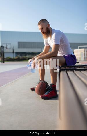 Joueur de basket-ball à barbe avec eau et ballon assis sur le banc et reposant sur le terrain Banque D'Images