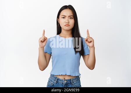 Une femme asiatique en colère brune pointant les doigts vers le haut, frognant et regardant déçu de la caméra, debout dans un t-shirt bleu sur fond blanc Banque D'Images