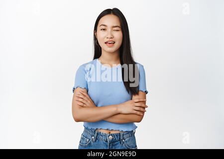 Sassy brunette asiatique fille wencing à la caméra et sourire, vous assurer, en faisant allusion à quelque chose, debout avec les bras croisés sur fond blanc Banque D'Images