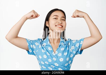 Gros plan sur une femme asiatique souriante, qui se sent forte et puissante, qui fléchit les biceps et qui semble fière, montrant ses muscles forts, se tenant sur du blanc Banque D'Images