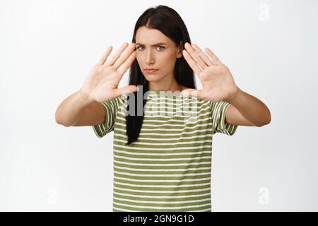 Femme sérieuse étire les mains dans le bloc, s'arrêtant, disons non, frognant et regardant déplu, debout sur fond blanc Banque D'Images